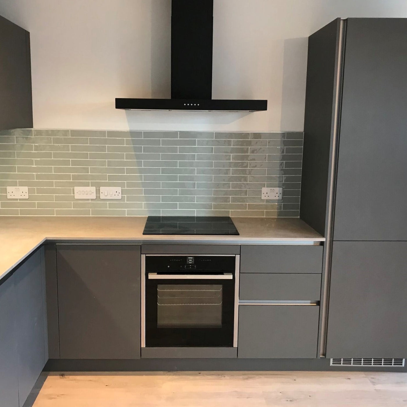 Black FLT wall mounted cooker hood installed in a modern grey kitchen , tiled surround and light grey countertop 